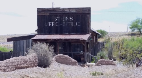 Abandoned wooden mercantile building with a faded sign, surrounded by desert landscape and sparse vegetation.