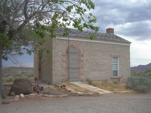 A small stone building with a gray door, surrounded by sparse vegetation and a tree, under a cloudy sky.