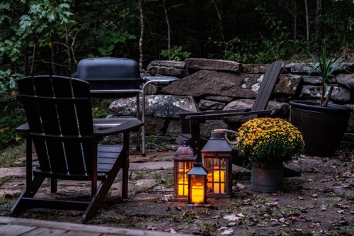 Cozy outdoor seating area with lantern, flowers, and a grill, surrounded by stone walls and trees at dusk.