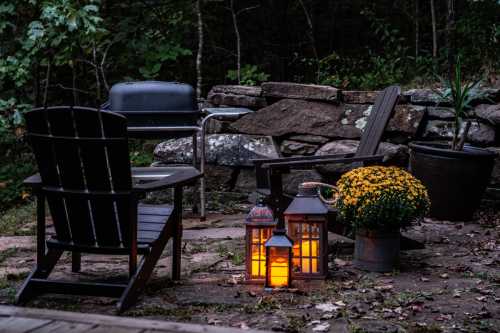 Cozy outdoor seating area with lantern, flowers, and a grill, surrounded by stone walls and trees at dusk.