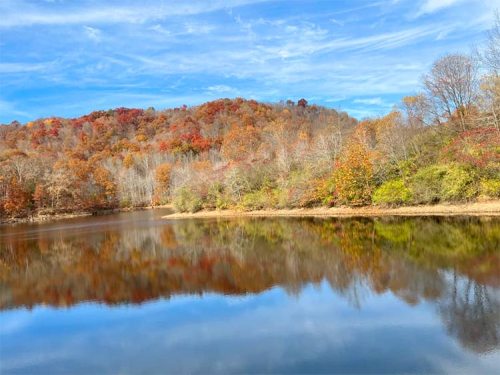 A serene lake reflecting colorful autumn foliage and a clear blue sky, surrounded by trees on a calm day.