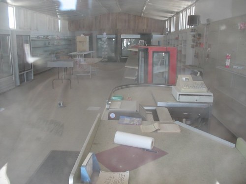 An empty, abandoned store interior with shelves, a counter, and scattered items, illuminated by natural light.