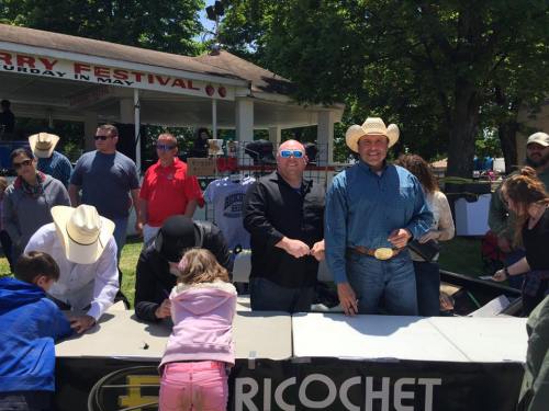 A lively outdoor festival scene with people gathered around tables, some wearing cowboy hats and sunglasses.