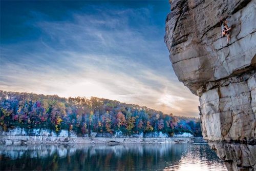A climber ascends a rocky cliff by a calm lake, surrounded by colorful autumn foliage and a scenic sky.