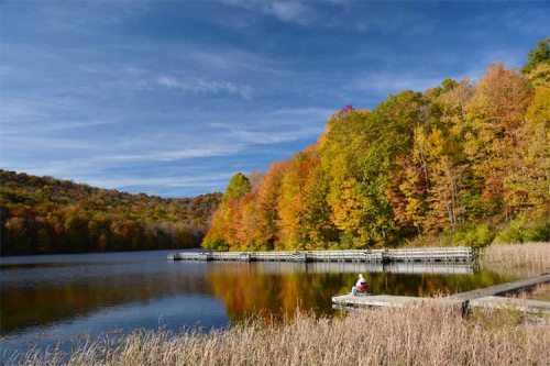 A serene lake surrounded by vibrant autumn foliage, with a person sitting on a dock enjoying the view.