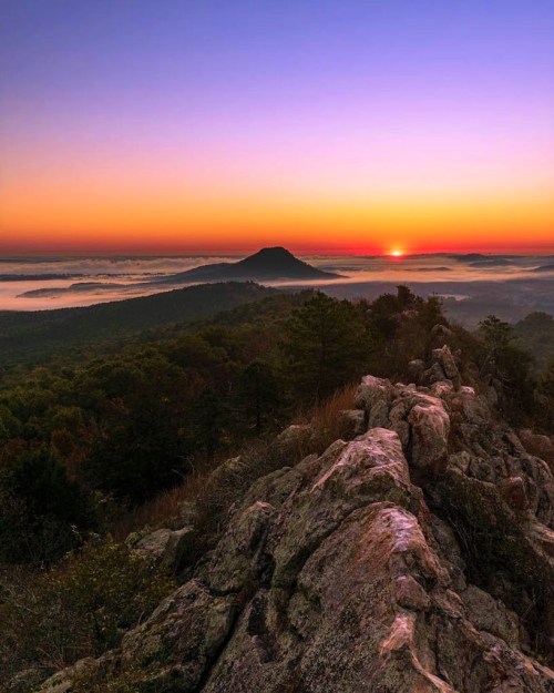 A stunning sunrise over misty mountains, with rocky foreground and vibrant orange and purple sky.