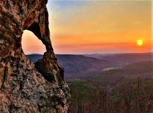 A rocky outcrop frames a sunset over rolling hills, casting warm colors across the sky and landscape.