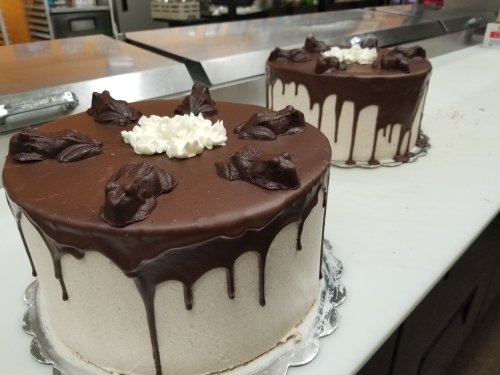 Two chocolate-drizzled cakes with decorative frosting on top, displayed on a countertop in a bakery.