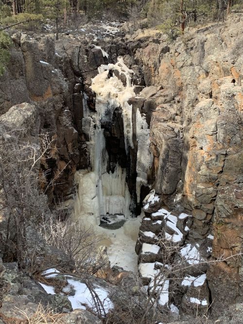 A frozen waterfall cascading down rocky cliffs, surrounded by snow and trees in a rugged landscape.