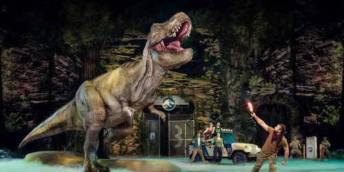 A lifelike T. rex roars on stage while a performer holds a flare, surrounded by a jungle setting and a jeep.