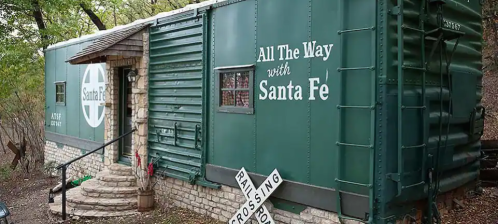 A green train car converted into a cozy cabin, featuring a stone entrance and a sign that reads "All The Way with Santa Fe."