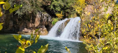 A serene waterfall cascades into a clear pool, surrounded by lush greenery and autumn foliage.