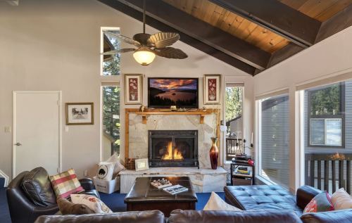 Cozy living room with a stone fireplace, large windows, and modern decor, featuring a ceiling fan and comfortable seating.