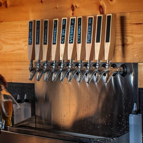 A row of beer taps with wooden handles, labeled with different beer styles, against a wooden wall.