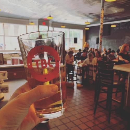 A hand holds a glass of beer with a logo, set in a lively bar with patrons in the background.
