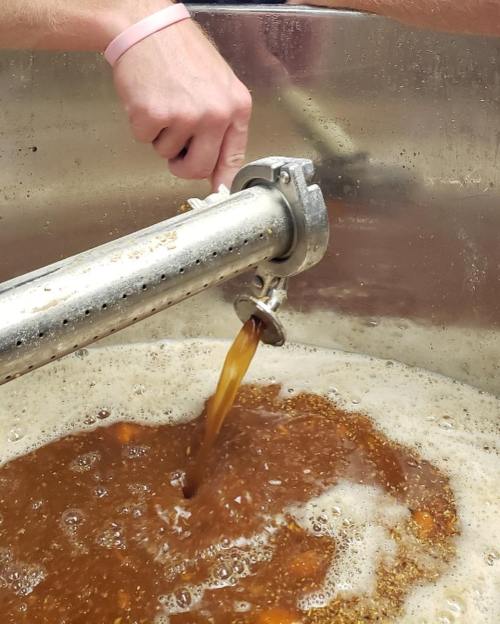 A hand adjusts a valve on a brewing tank, with brown liquid bubbling inside.
