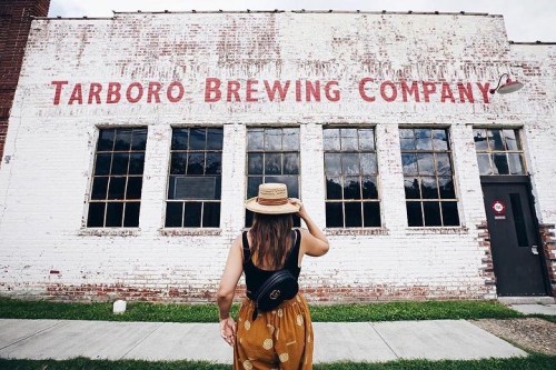 A woman in a straw hat stands in front of the Tarboro Brewing Company building, admiring its brick facade.