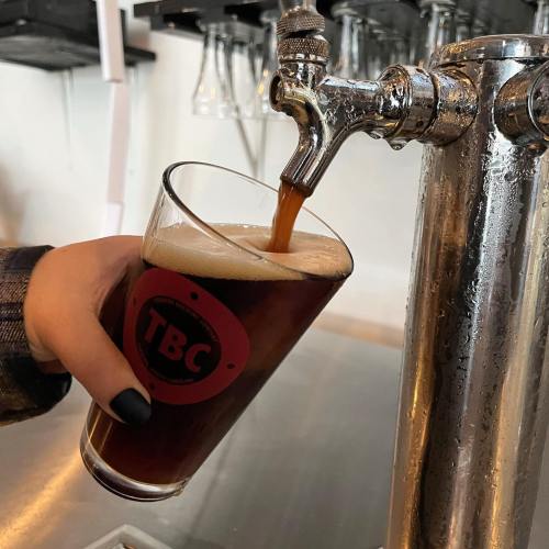 A hand holds a glass as dark beer pours from a tap into it, with a brewery logo visible on the glass.