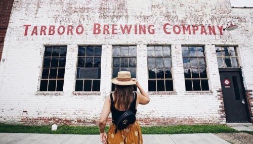 A woman in a straw hat stands in front of the Tarboro Brewing Company building, looking at the sign.