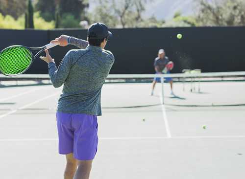 A man in a gray shirt and purple shorts hits a tennis ball while another player prepares to return it on a court.