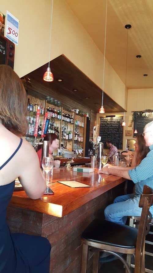 A cozy bar scene with patrons seated at the counter, a bartender preparing drinks, and shelves of liquor in the background.