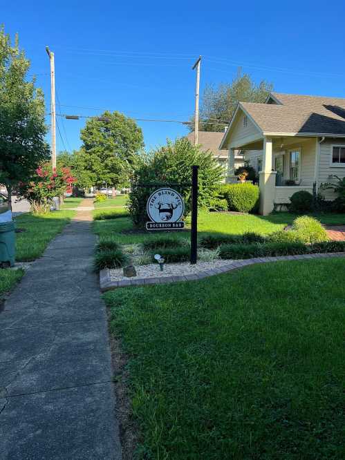 A pathway leads to a house with a sign for "Blind Bourbon Rd" surrounded by green grass and trees under a clear blue sky.