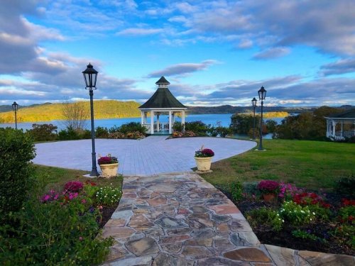 A scenic view of a gazebo by a lake, surrounded by flowers and lampposts under a blue sky with clouds.