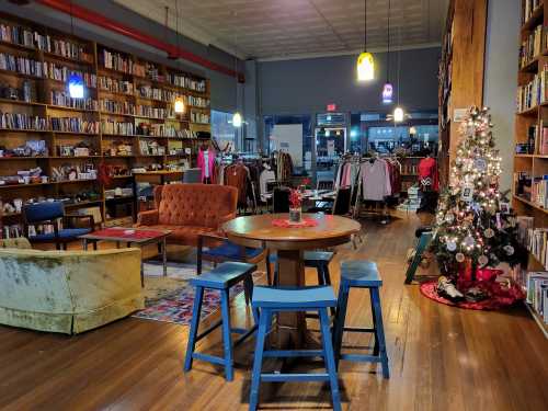 Cozy bookstore with bookshelves, a decorated Christmas tree, seating areas, and clothing racks in a warm, inviting atmosphere.