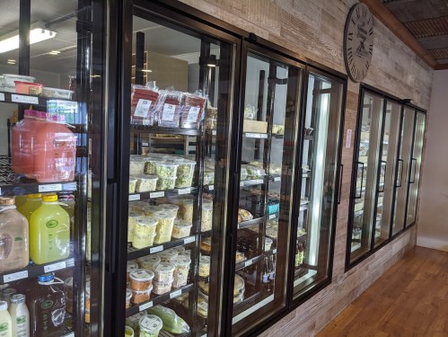 Refrigerated display cases filled with various packaged food items in a store. Wooden wall and clock in the background.