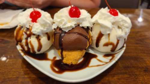 Three ice cream scoops topped with whipped cream and cherries, drizzled with chocolate sauce on a plate.