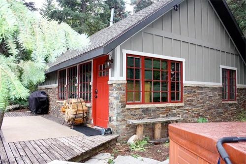 A cozy cabin with a stone exterior, red door, and large windows, surrounded by trees and a wooden deck.