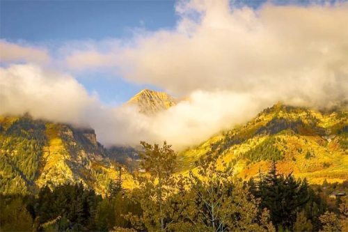A scenic mountain landscape with autumn foliage, partially obscured by clouds under a blue sky.