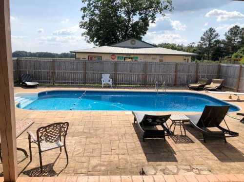 A backyard pool area with a blue pool, lounge chairs, and a wooden fence under a sunny sky.