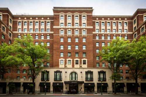 A large brick building with multiple windows and green trees in front, showcasing a classic architectural style.