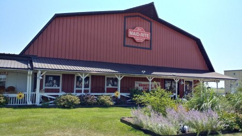 A red barn-style building with a "Maid-Rite" sign, surrounded by green grass and colorful flowers.