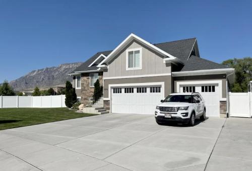 A modern house with a two-car garage, parked white SUV, and a mountain backdrop under a clear blue sky.