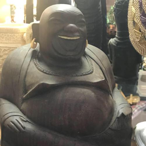 A smiling wooden statue of a Buddha figure, featuring a round belly and large earrings, surrounded by other decorative items.