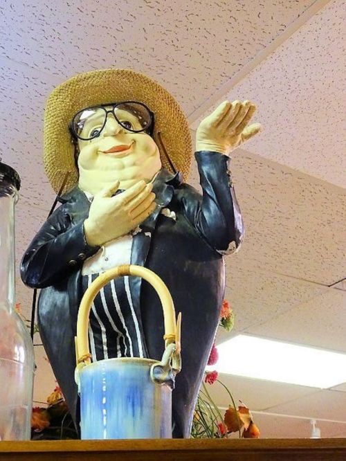 A whimsical ceramic figure of a smiling man in a suit and straw hat, waving with one hand, atop a shelf.