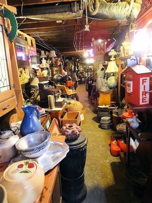 A cluttered antique shop aisle filled with various vintage items, lamps, and collectibles under warm lighting.