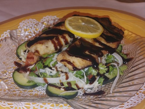 A plated dish featuring grilled fish topped with a lemon slice, served on a bed of fresh vegetables and drizzled with sauce.