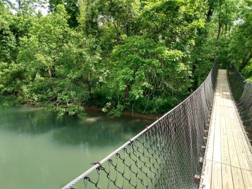 A wooden suspension bridge spans a calm river, surrounded by lush green trees and foliage.