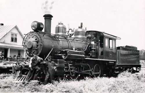 A vintage steam locomotive with two men standing beside it, set against a backdrop of a house and grassy area.