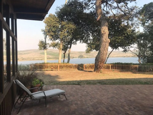 A serene lakeside view from a porch, featuring trees and a wooden fence in the foreground.
