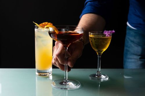 A hand reaches for a cocktail, with two other drinks on a glass surface: a tall orange drink and a small floral garnish drink.