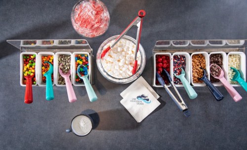 A variety of toppings including candies, nuts, and marshmallows arranged in containers, with scoops and a bowl in the center.