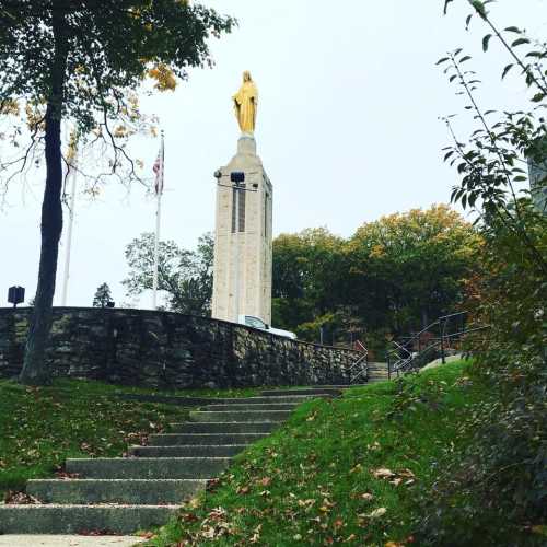 A statue atop a stone monument surrounded by trees, with steps leading up to it on a cloudy day.