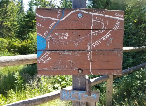 Wooden trail sign with a map indicating trails and "You Are Here" location in a forested area.