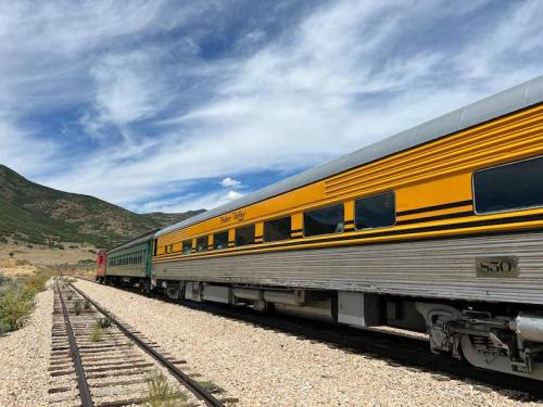A vintage train with a yellow and green car parked on a gravel track, surrounded by mountains and a blue sky.