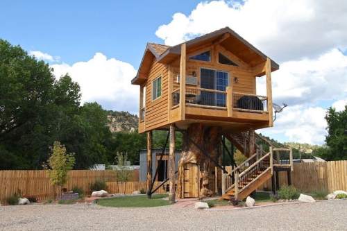 A charming wooden treehouse with a balcony, set in a scenic outdoor area surrounded by trees and a wooden fence.