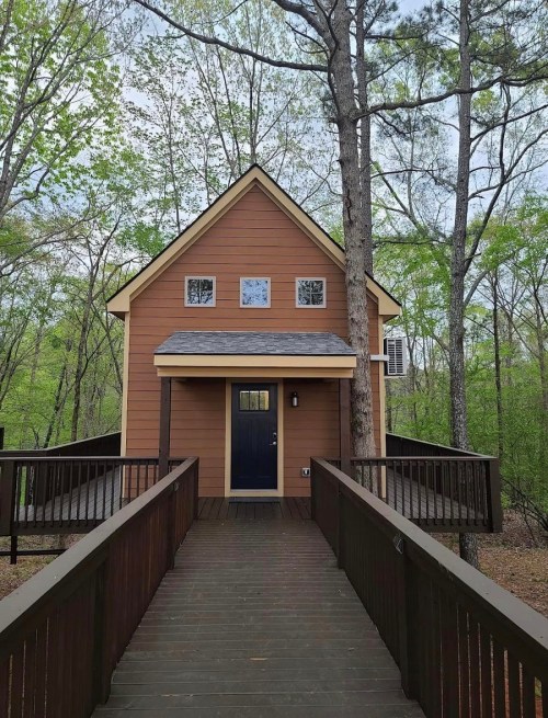 A small, brown cabin with a sloped roof, surrounded by trees, accessed by a wooden walkway.